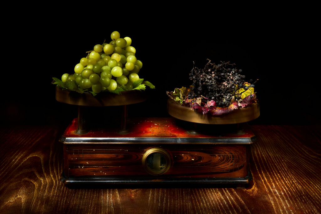 Fotografia fine art pittorialista "La bilancia del tempo" di Antonio Milesi: natura morta Vanitas con bilancia antica che pesa uva fresca e uva appassita, stile chiaroscuro ispirato al Romanticismo. Fine art pictorialist photograph "The Scales of the Ages" by Antonio Milesi: Vanitas still life with an antique scale balancing fresh grapes and withered grapes, chiaroscuro style inspired by Romanticism.