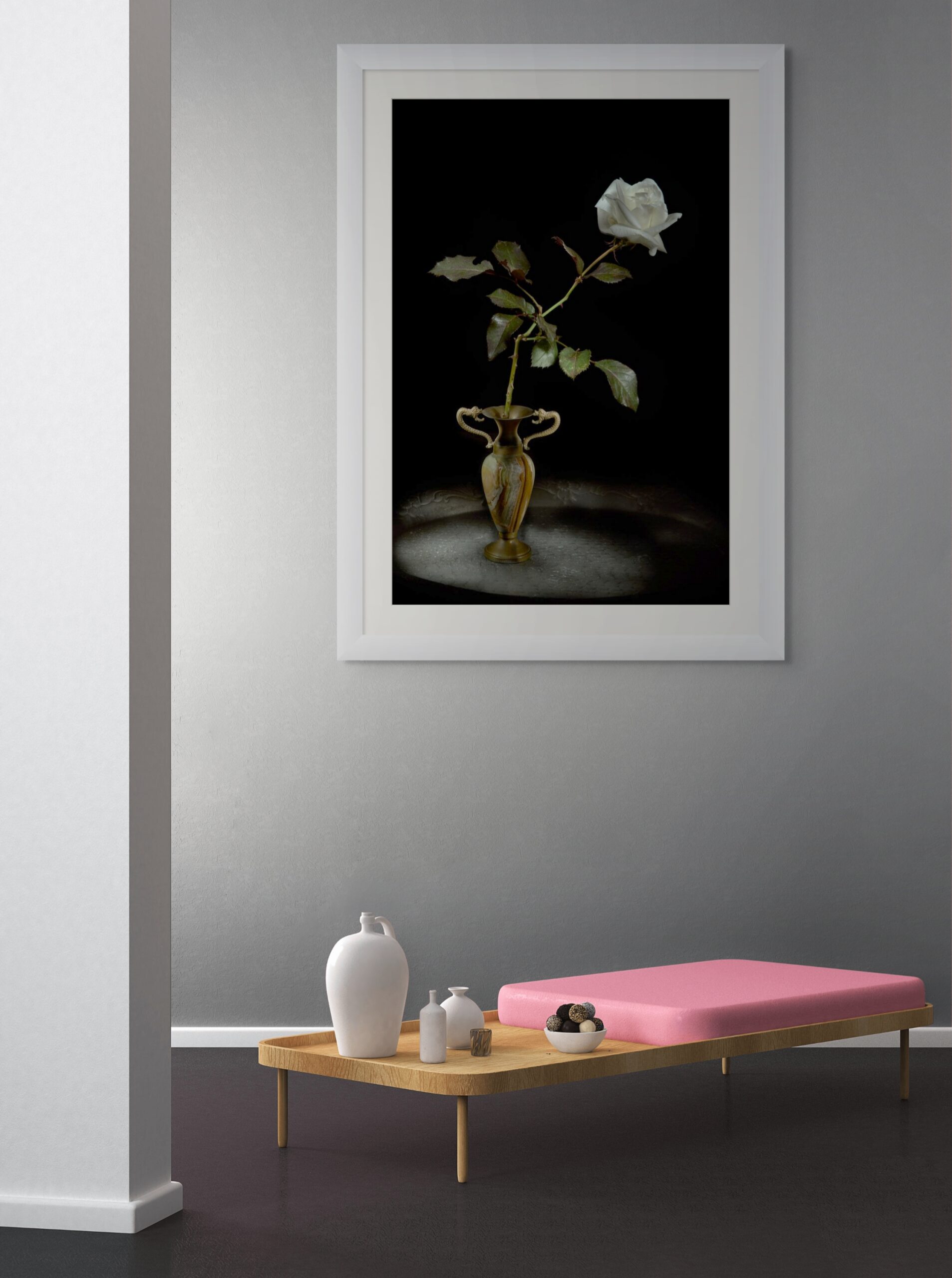 Mockup di interni minimalista con l'opera "Elegia della Luce Bianca" di Antonio Milesi. La fotografia fine art, con una rosa bianca e vaso ornamentale su sfondo nero, è esposta in una stanza luminosa e moderna con pareti grigie e arredi essenziali. Minimalist interior design mockup featuring "Elegy of White Light" by Antonio Milesi. The fine art photograph, showing a white rose and ornamental vase against a black background, is displayed in a bright modern room with grey walls and essential furniture.